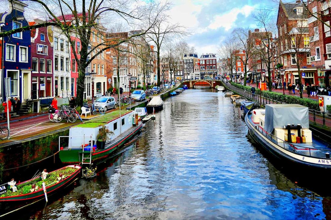 Houseboats moored along a narrow Amsterdam canal lined with colourful brick houses, parked bikes and bare winter trees