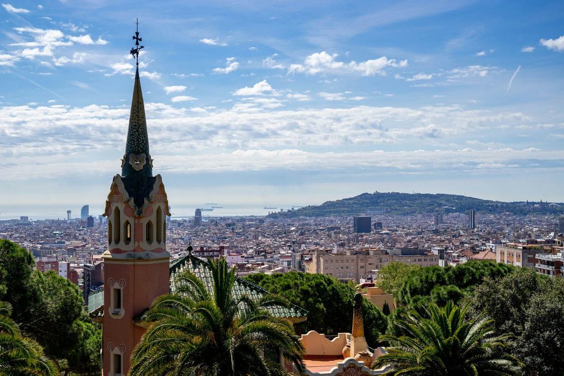 Barcelona from Park Güell — the spire of the Gaudí House Museum and palm trees in front, the city and Montjuïc hill beyond