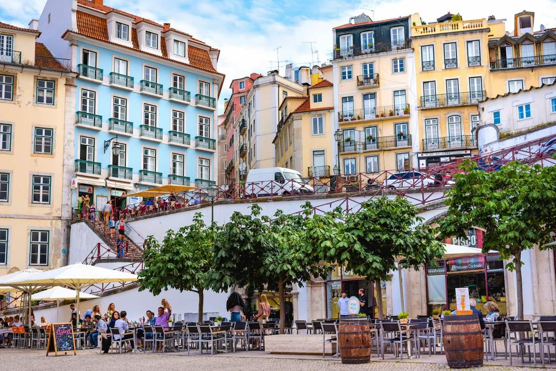 Pastel-coloured Lisbon buildings with iron balconies above a busy square of café terraces and trees
