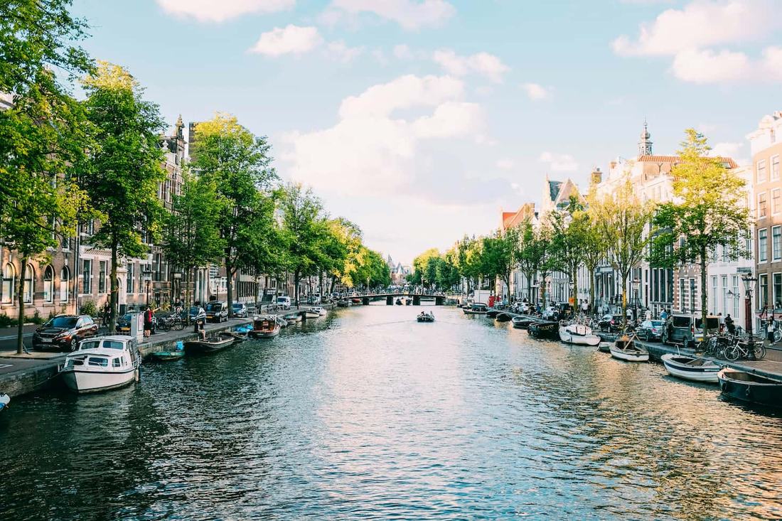 A tree-lined Amsterdam canal with moored boats and gabled canal houses, a bridge in the distance
