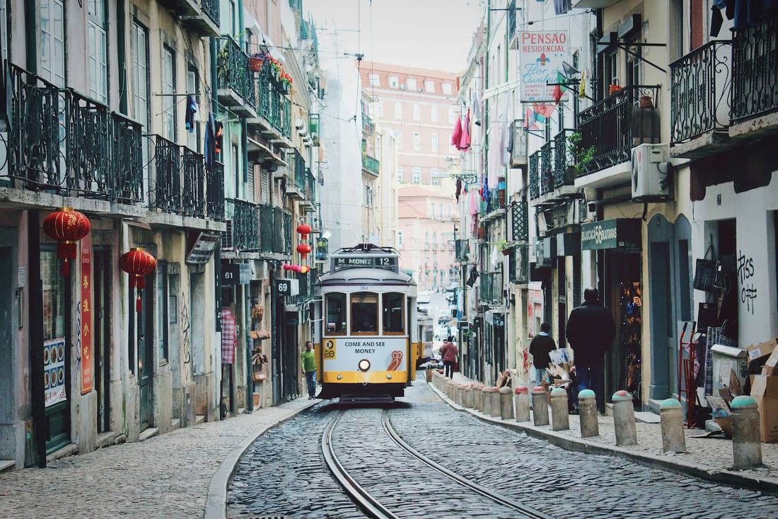 A yellow vintage tram on a narrow cobbled street in Lisbon, lined with iron balconies and shop signs
